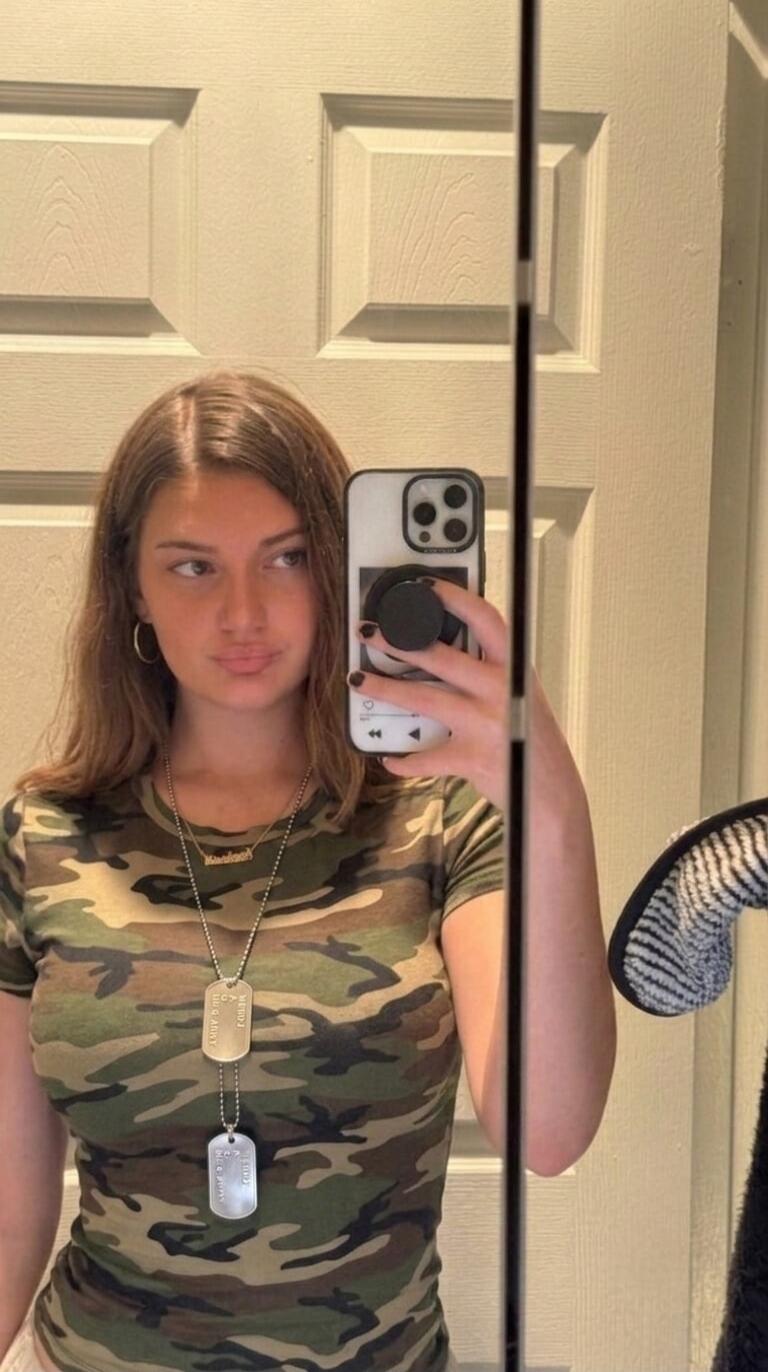 A young woman takes a mirror selfie, wearing a camouflage t-shirt and two dog tags. One dog tag appears to say 'U.S. ARMY' and 'MERCH'. She is holding a smartphone with a pop socket and a phone case designed to look like an iPod classic. She has a small necklace with a name, possibly 'Kashish'.
