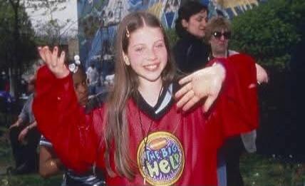 A young girl in a red jersey with a circular logo posing outdoors.