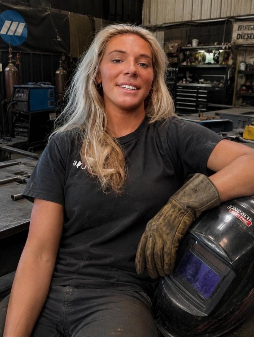 A young blonde woman in a workshop, wearing a dark t-shirt and work pants, with a work glove on her right hand, leaning on a Lincoln Electric welding helmet. She is smiling at the camera. In the background, a Miller logo and a sign with 'DIESEL' are visible, along with welding equipment and gas cylinders.