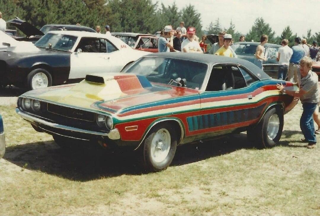 A vintage Dodge Challenger with a distinctive multi-colored stripe paint job and a large hood scoop is being pushed by a man on a grassy field. Other classic cars and people are visible in the background, suggesting a car show or event.