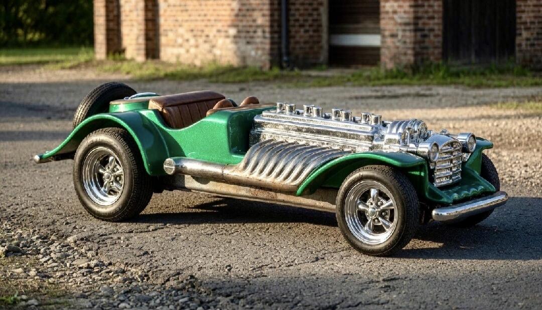 A custom-built, green vintage-style hot rod or custom car with a prominent chrome engine and side exhaust pipes, parked on a gravel road.