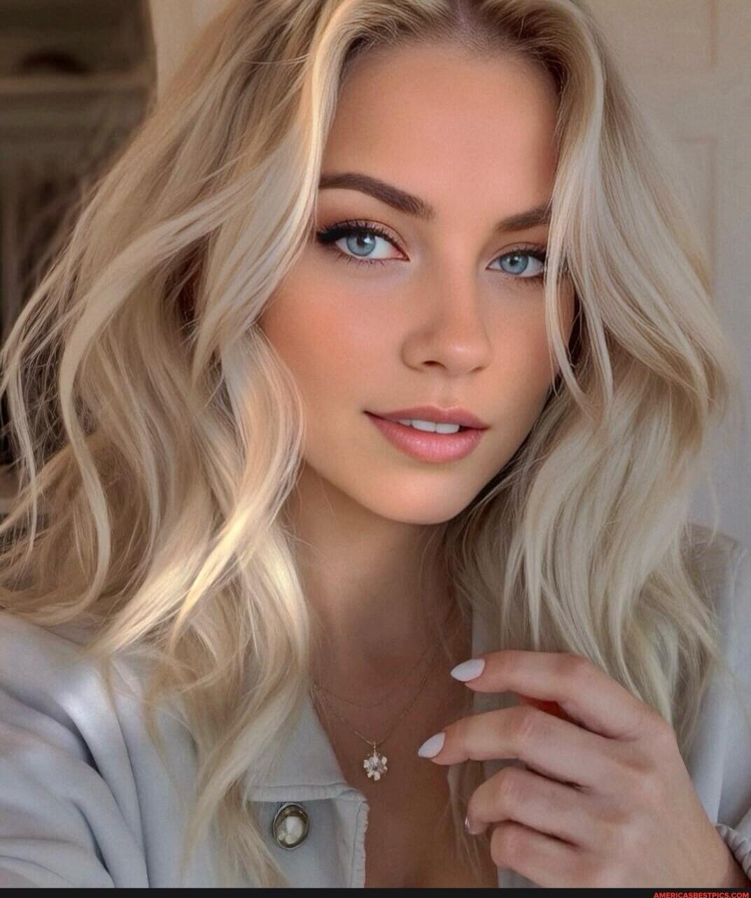 A close-up portrait of a young woman with long, wavy blonde hair and blue eyes. She has light makeup, a soft pink lip, and is wearing a light-colored jacket. She is touching a delicate necklace with a small pendant.