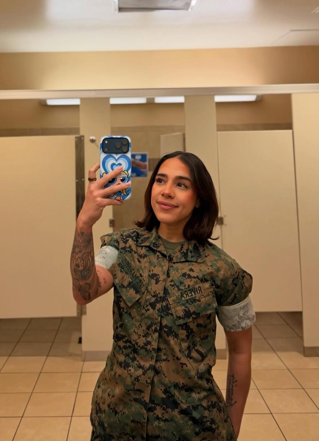 A woman in a military uniform taking a mirror selfie in a restroom. She has tattoos on her arms, some covered with white sleeves. Her uniform has the name 'RIVERA' (mirrored) on it. She is holding a phone with a blue heart case.