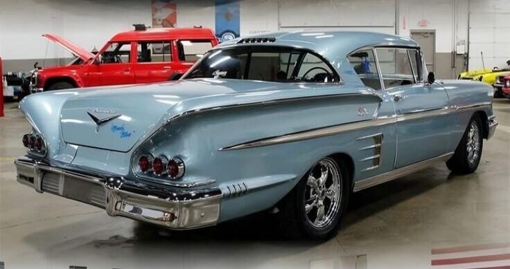 A light blue vintage car, possibly a Chevrolet Impala, with custom chrome wheels, parked in a garage or showroom. A red truck with an open hood is visible in the background. Text on the car's trunk appears to be 'Impala SS'.