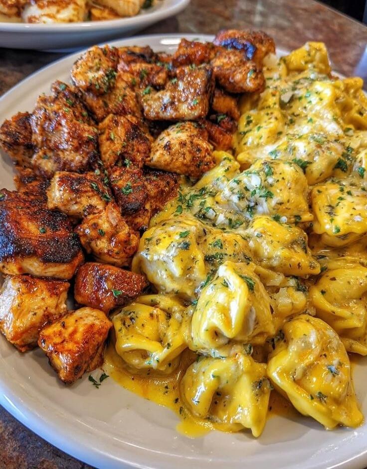 A plate featuring two distinct sections: on the left, browned, crispy cubed meat (likely grilled or fried), and on the right, yellow cheese-tortellini or pasta covered in a rich cheese sauce with herbs.