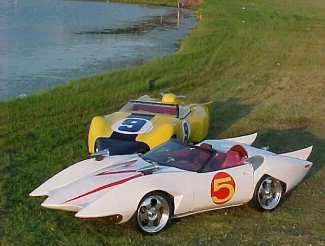 White vintage race car with red stripe and number 5 beside a yellow vintage race car with blue circle and number 9, parked on grassy bank near a small body of water.