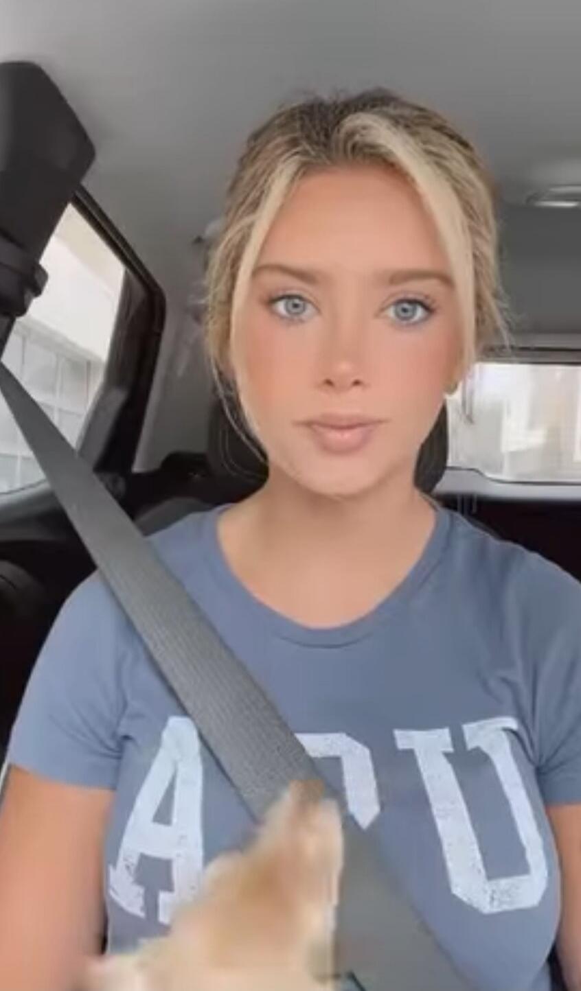 A young woman with blonde hair and blue eyes is sitting in a car, wearing a blue t-shirt and a seatbelt. A small pet is partially visible in the foreground.