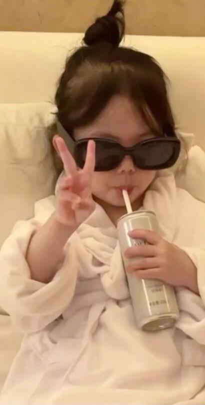 A young child wearing sunglasses, sipping from a bottle with a straw and making a peace sign.