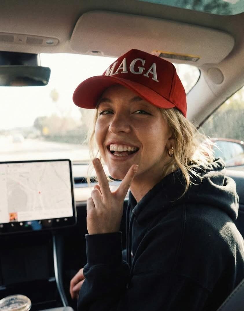 A young woman with blonde hair, wearing a red cap with 'MAGA' written on it and a black hoodie, is smiling broadly and making a peace sign with her fingers while sitting in a car. A navigation map is visible on the car's central screen.