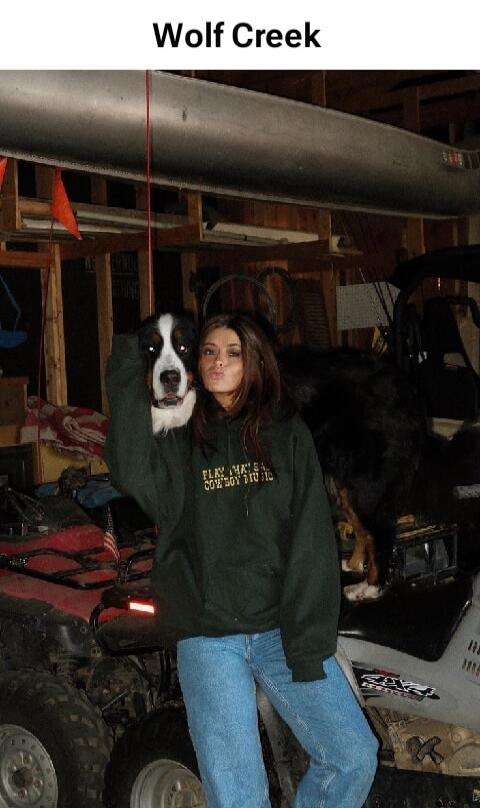 Wolf Creek. A woman in a green hoodie and blue jeans is posing with a Bernese Mountain Dog, holding its head next to her face and making a kissing expression. The hoodie has text that appears to say 'PLAY MA'S COWBOY STUDIO'. In the background, there are ATVs and a canoe hanging overhead.
