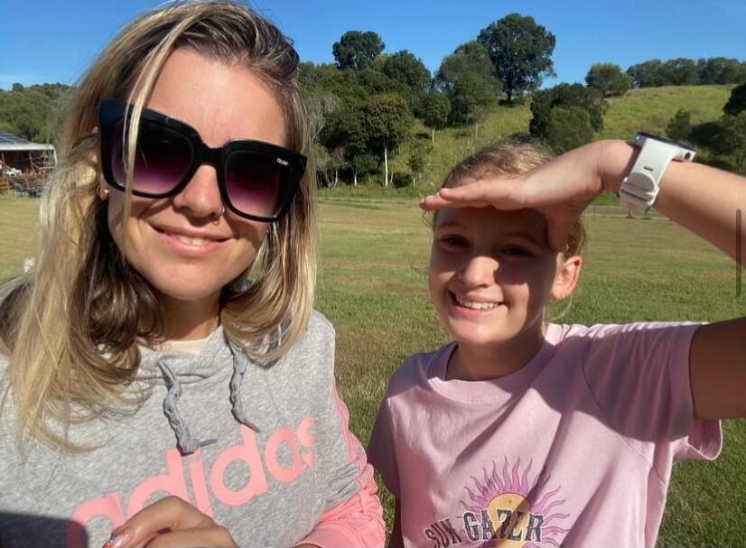 Two people posing outdoors on a sunny day: a woman wearing sunglasses and a gray Adidas hoodie, and a child in a pink shirt shielding their eyes from the sun.
