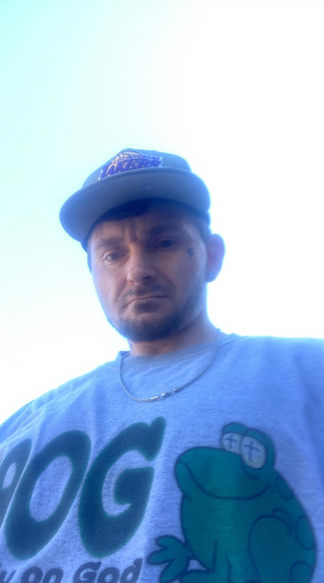 A man wearing a Lakers cap and a t-shirt with 'OG on God' and a frog cartoon. The man has a small tattoo near his eye.