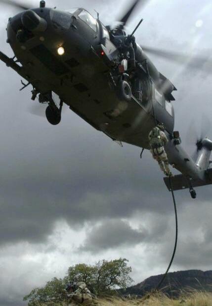 A military helicopter is hovering while a soldier fast-ropes down from it. Another soldier is on the ground, possibly securing the area or waiting for the descent.