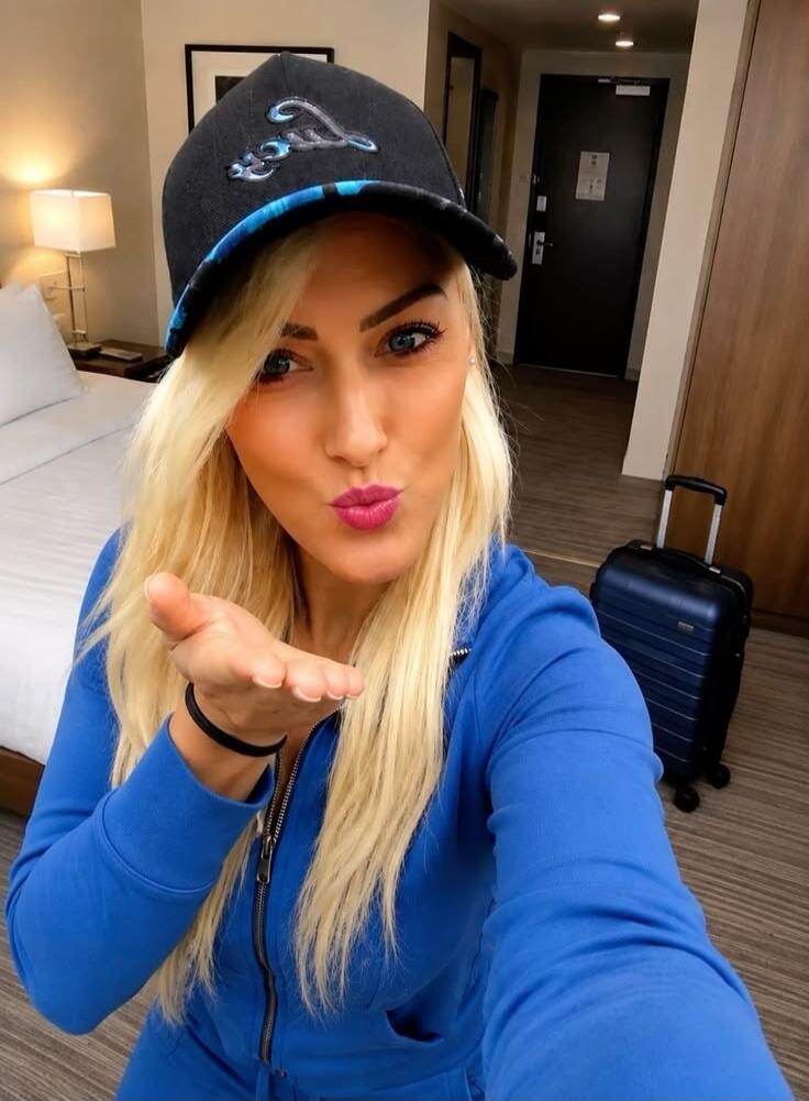 A blonde woman in a blue tracksuit and a black cap is taking a selfie in a hotel room, making a kiss face and blowing a kiss towards the camera.