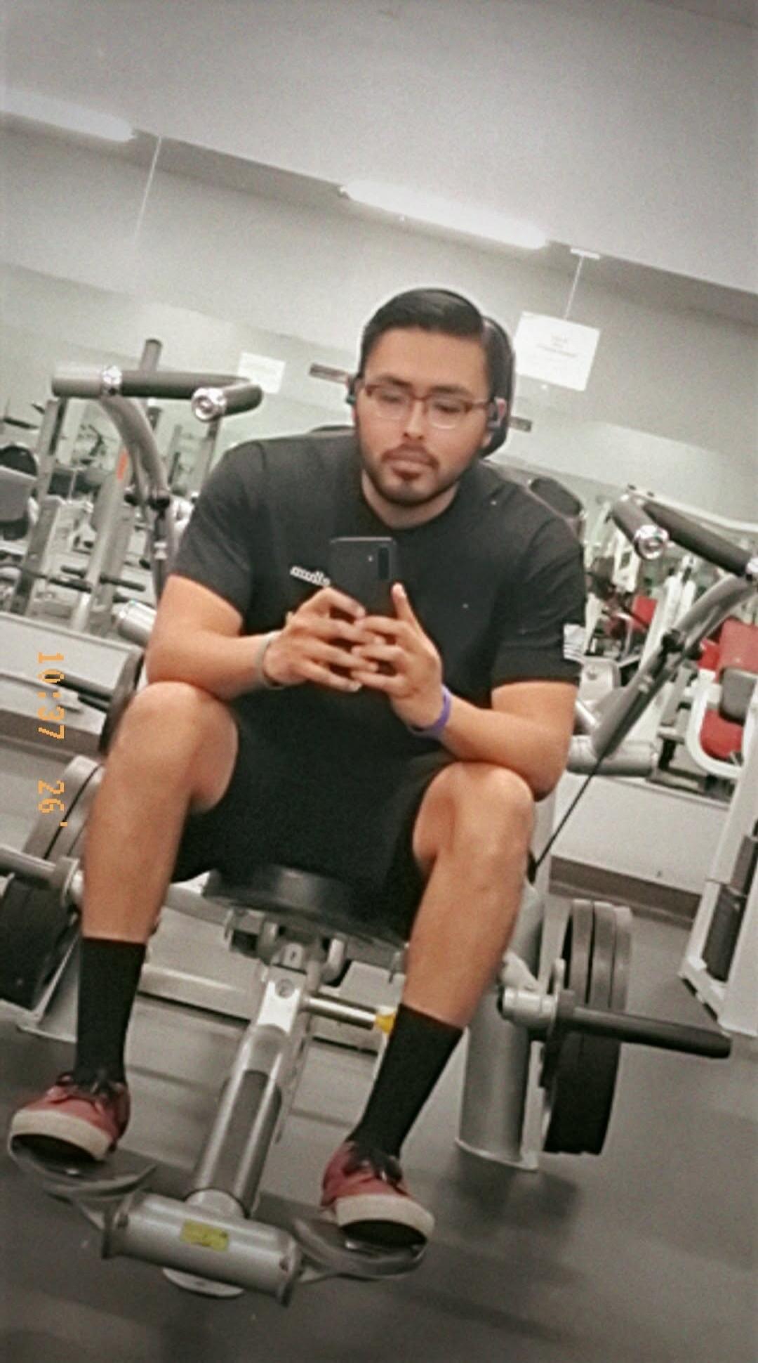 10 7 26. A man is sitting on a gym machine, taking a selfie with his phone. He is wearing a black t-shirt, black shorts, black socks, and red sneakers. He has glasses and headphones. The background shows other gym equipment and mirrors.