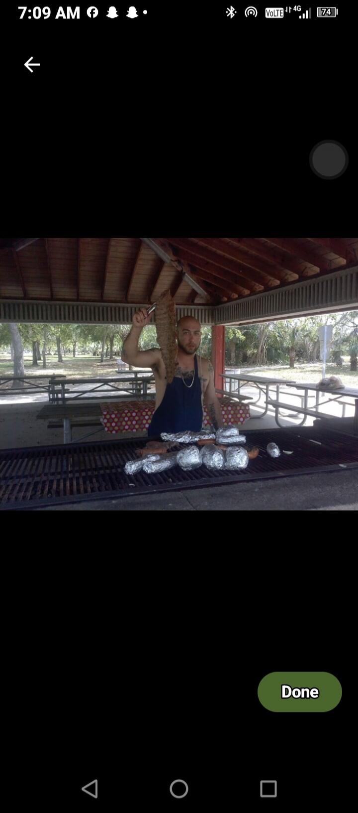 A man is grilling a large piece of meat and other food items wrapped in foil on a barbecue grill in an outdoor park setting.