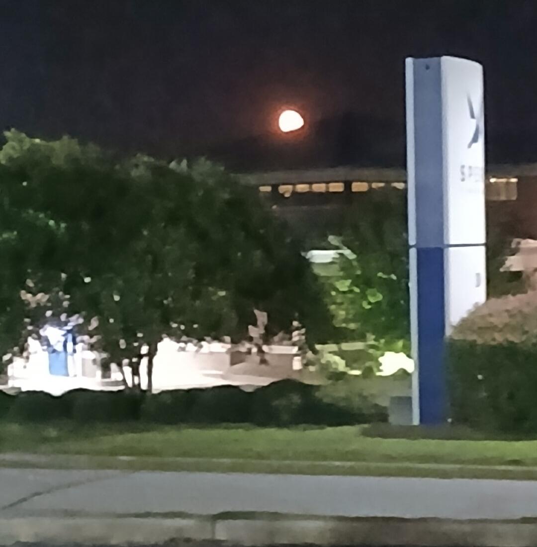 A reddish-orange half-moon in a dark night sky, with trees and a building in the background. A white and blue signpost with the text 'SPI' stands to the right. Grass and a road are in the foreground.