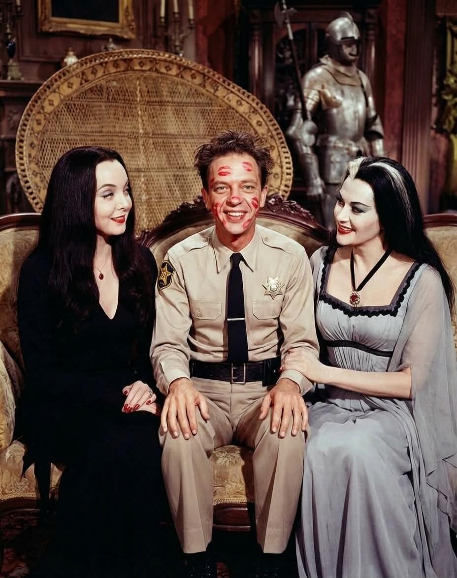 A man in a sheriff's uniform with his face covered in red lipstick kiss marks, sitting between two women dressed in gothic attire, resembling Morticia Addams and Lily Munster. All three are smiling.