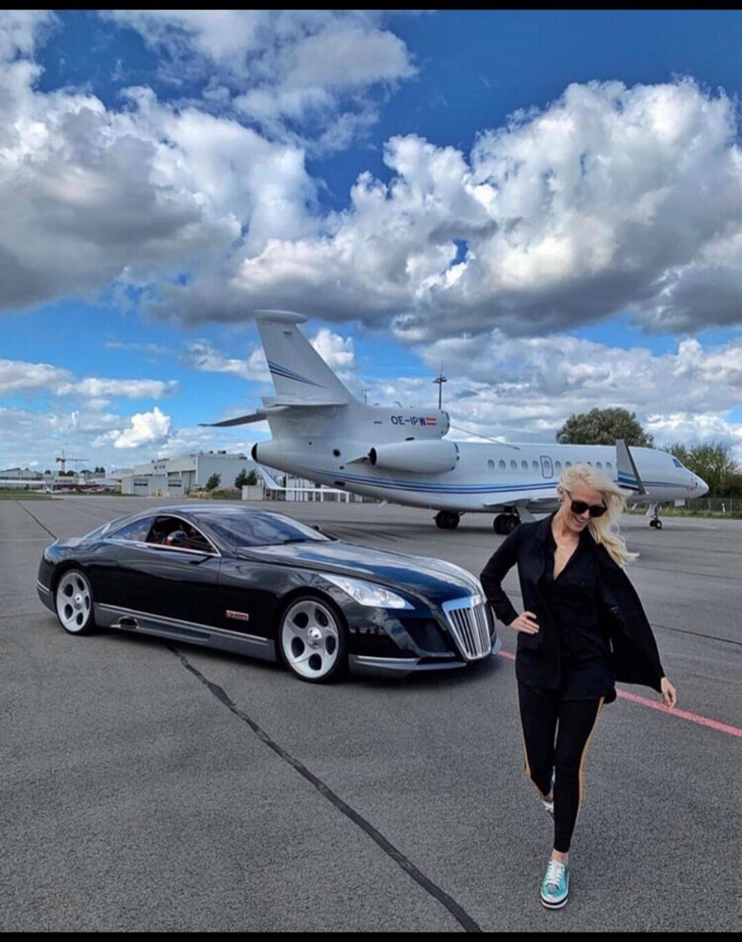 A woman walking on an airfield next to a black luxury car and a private jet with tail number OE-IPW.