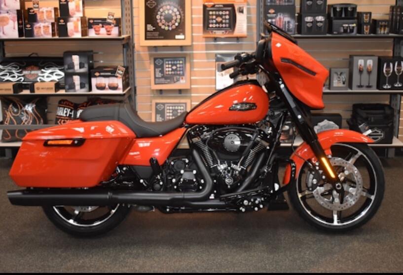 A vibrant orange Harley-Davidson motorcycle is displayed in a showroom, with various branded merchandise on shelves in the background.