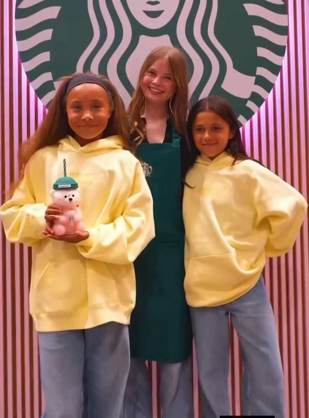 Three girls posing in front of a Starbucks logo. One girl is wearing a Starbucks apron, and two others are wearing matching yellow hoodies and jeans. One of them is holding a cute pink bear-shaped drink container.