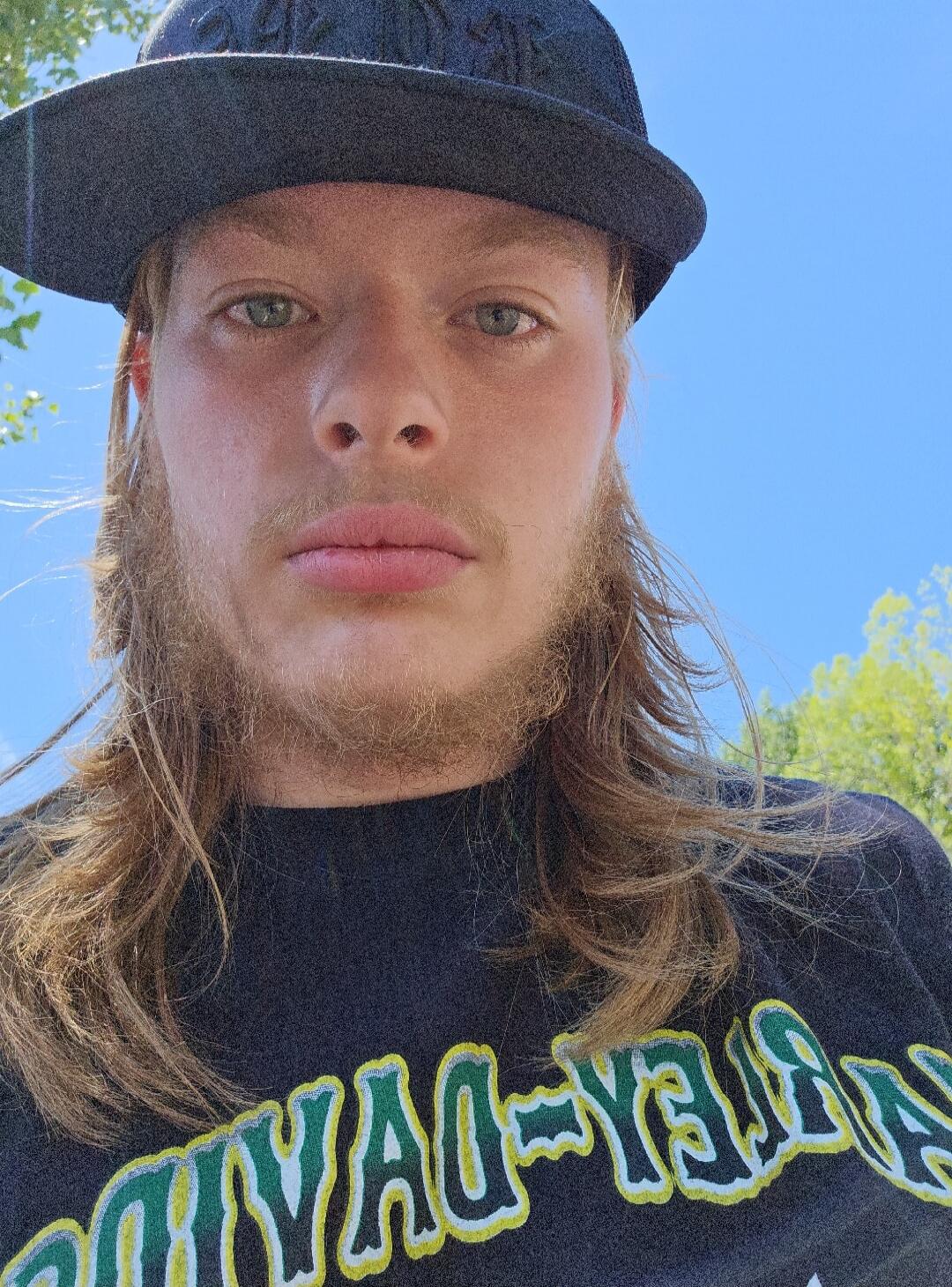 A close-up selfie of a young man with long blonde hair and a light beard, wearing a black hat and a dark t-shirt with partially visible text 'DAY-YAD'. The background shows a blue sky and green trees.