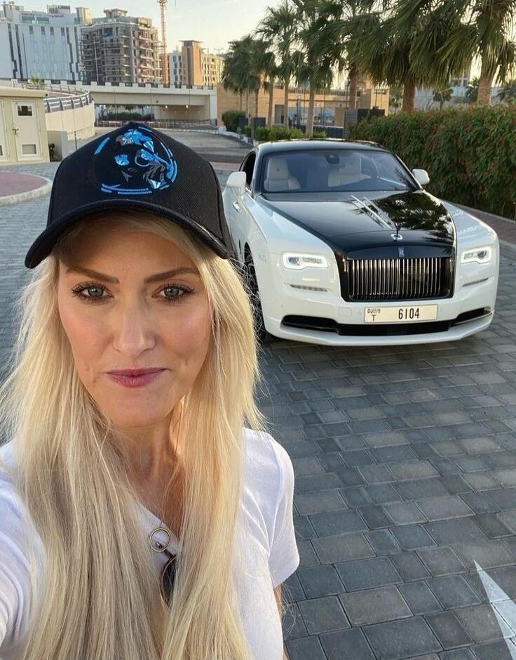 A blonde woman wearing a black cap with a blue panther logo and a white t-shirt takes a selfie. Behind her is a white and black Rolls-Royce car with the license plate 'DUBAI T 6104'. The background shows buildings and palm trees.