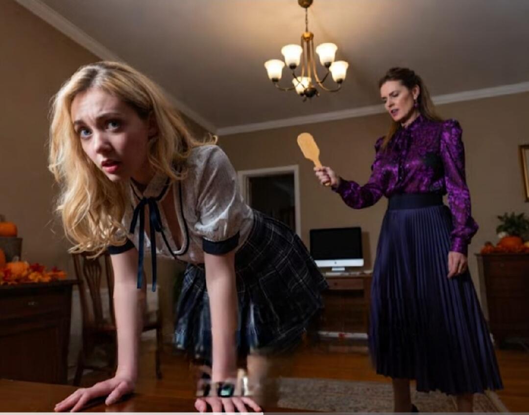 Two women in a living room: one is leaning over a table with a surprised expression, while the other stands behind holding a wooden paddle.