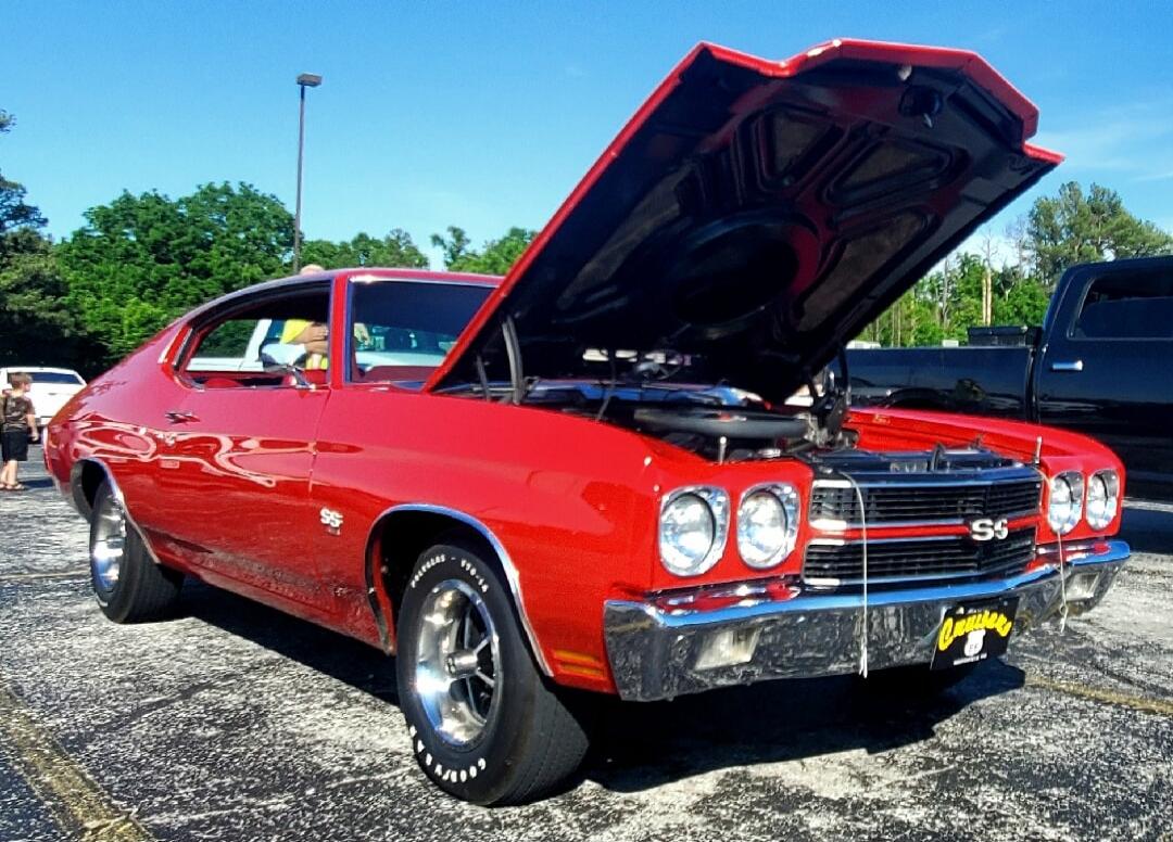 The image shows a red Chevrolet Chevelle SS with its hood open, revealing the engine. The car is parked outdoors on a sunny day, with a blue sky and trees in the background. There are other cars visible in the background, including a black pickup truck and a white car. A person is standing near the white car.