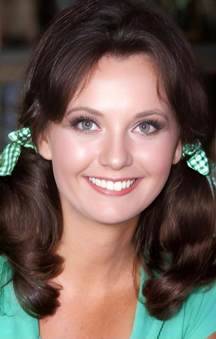 Beautiful young woman with brown hair styled in pigtails with green ribbons, wearing a teal top, and smiling at the camera.