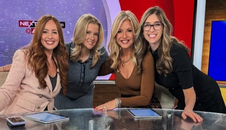 Four women posing at a news desk on a TV set.
