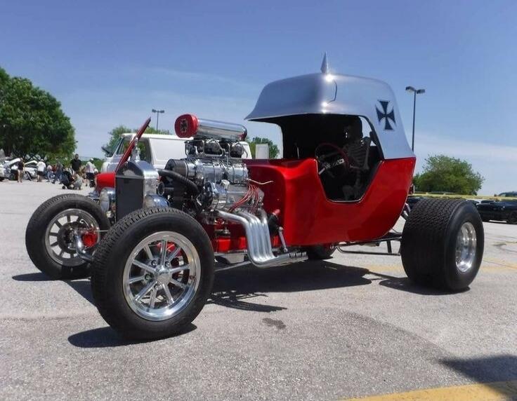 A custom-built hot rod car, possibly a T-bucket, with a bright red body and a unique silver roof shaped like a German Pickelhaube helmet, complete with an Iron Cross symbol on the side. The car features a large, exposed engine with a supercharger and chrome exhaust pipes. A skeleton figure is seated in the driver's seat. The vehicle is parked on as