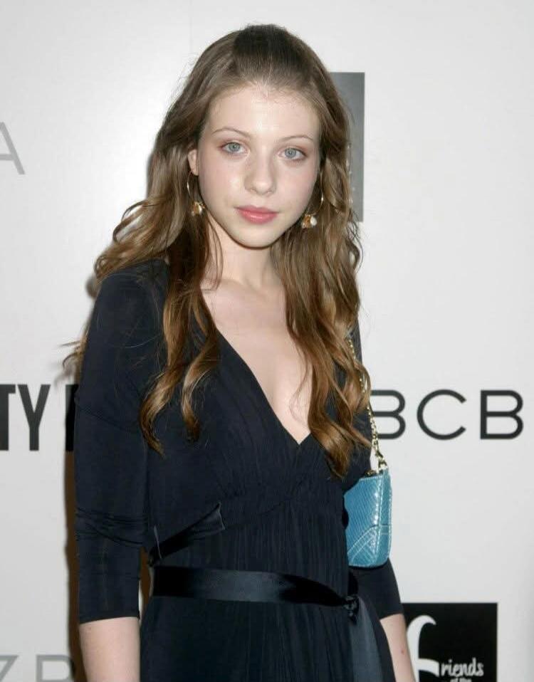 A young woman with long brown hair and blue eyes, wearing a black dress with a deep V-neckline and a black belt, holding a small blue purse. Partial text 'BCB' and 'friends of the' is visible in the background.