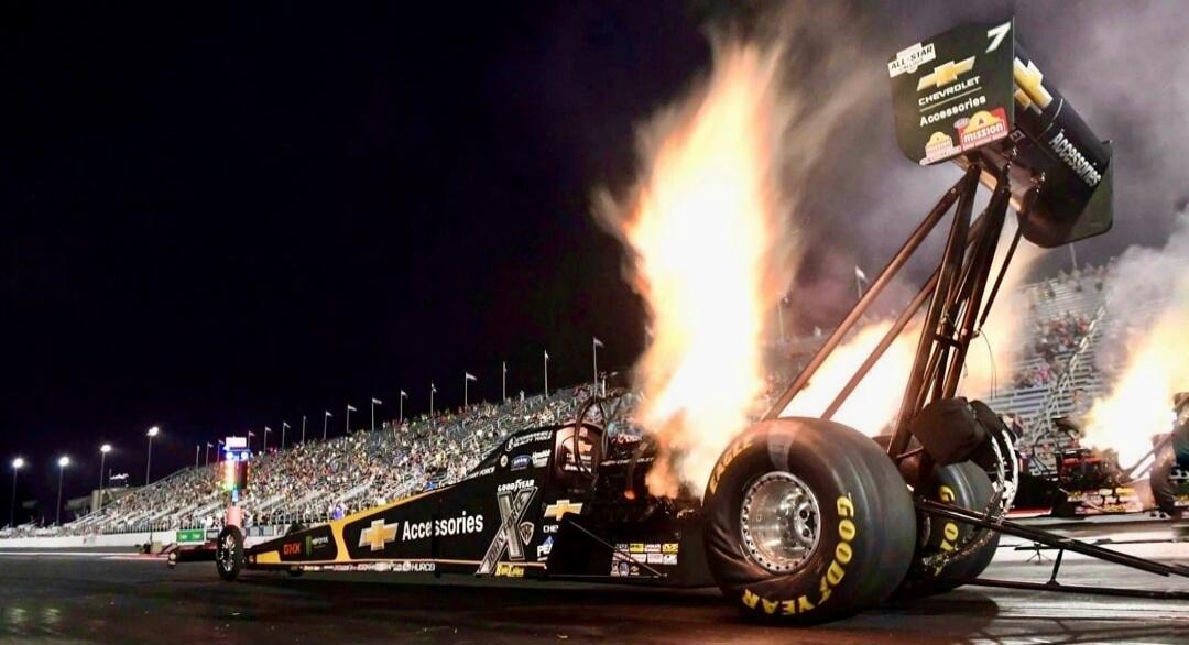 Goodyear on the tire; sponsor logos visible on the drag car; flames erupting from the exhaust; night race with large crowd stands in the background.