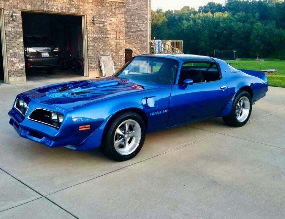 Blue vintage car (Pontiac Firebird Trans Am) parked on a driveway beside a brick building.