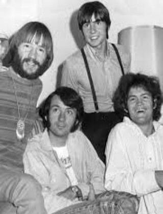A black and white photo of four men, possibly a band or group of friends, smiling and looking relaxed. They are casually dressed.