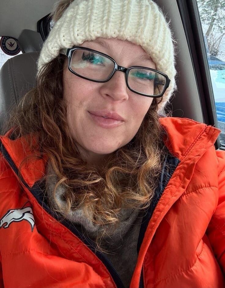 A woman wearing a white knit beanie and glasses sits in a car, dressed in an orange jacket over a dark top.