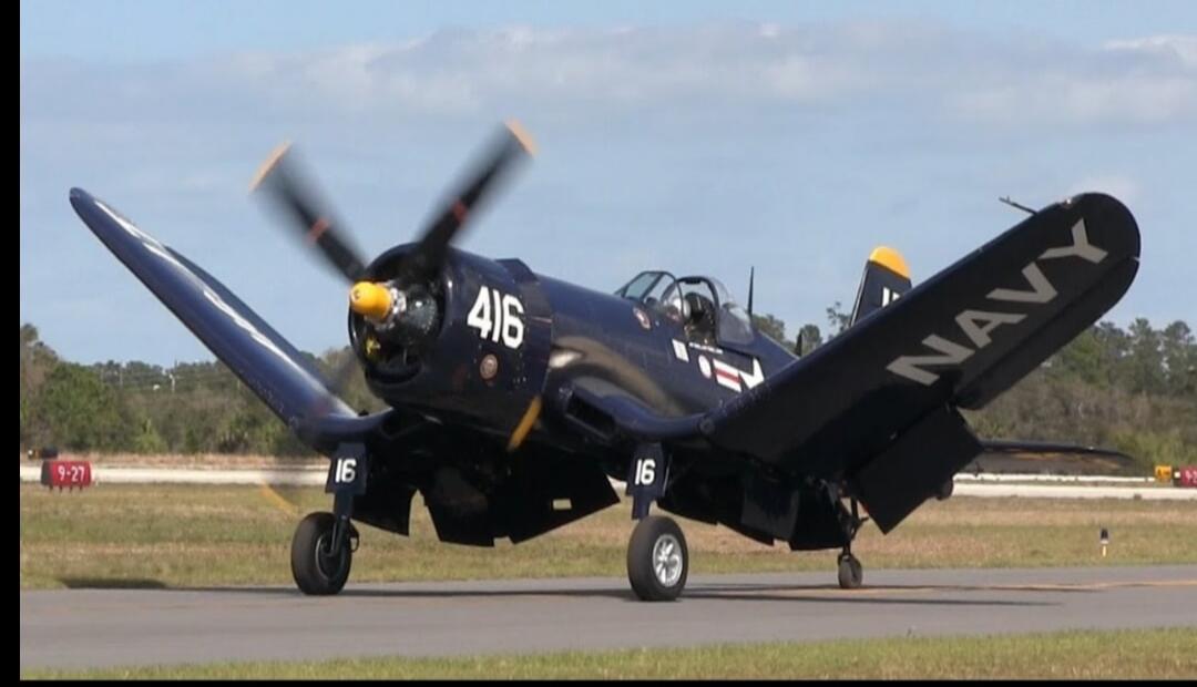 A dark blue vintage Vought F4U Corsair aircraft with '416' and 'NAVY' markings on its wings and fuselage, and '16' on its landing gear, is on an airfield. The propeller is spinning. A red sign in the background shows '9-27'.