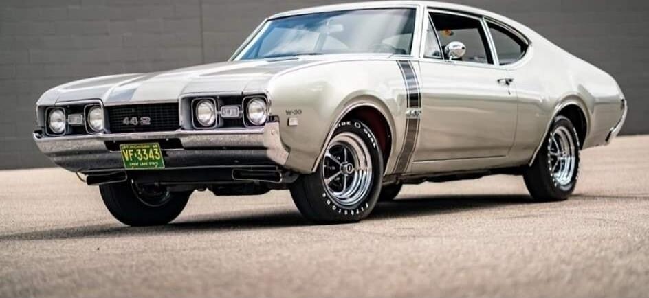 Beige classic muscle car parked on a paved surface. The car features a two-door coupe design with a prominent front grille, dual headlights, and chrome wheels. A green-yellow license plate is visible at the front.
