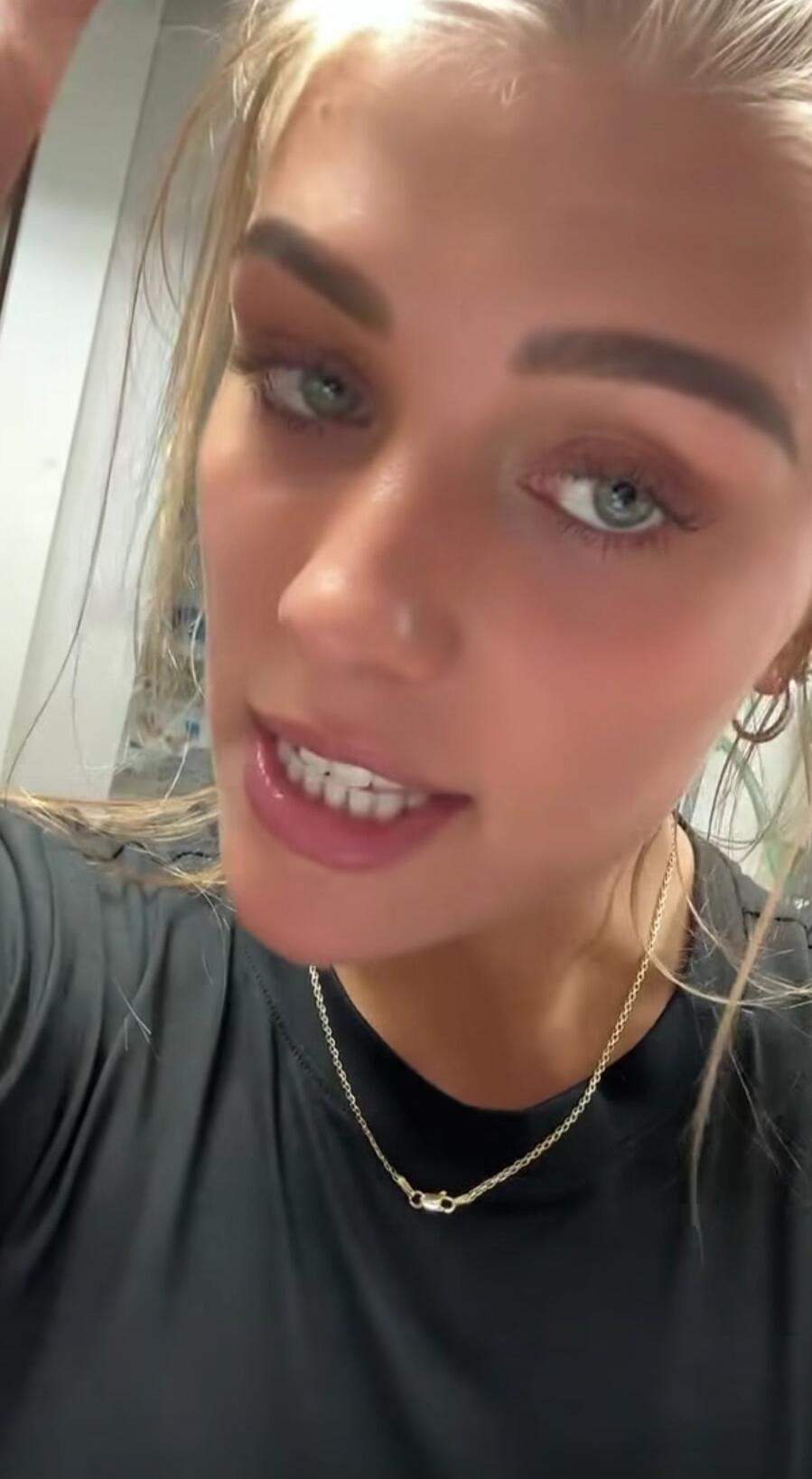 A young woman with blonde hair and light eyes is speaking directly to the camera, wearing a black top and a gold necklace.