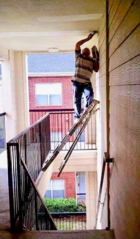 A man is painting a wall while standing on a ladder precariously balanced on a staircase railing, with one leg of the ladder extended over the void. The setup appears extremely unsafe.