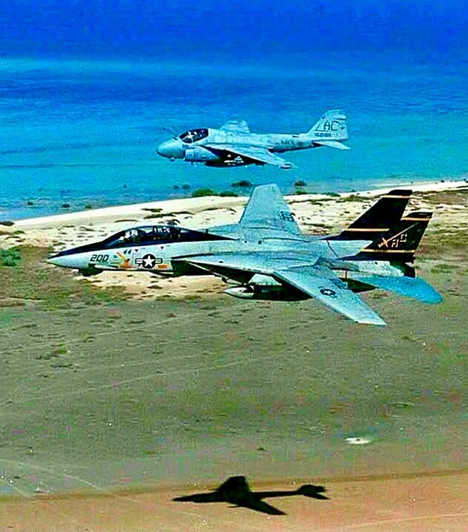 Two military jets, an F-14 Tomcat with '200' on its nose and an A-6 Intruder with 'AC' on its tail, flying over a coastline.