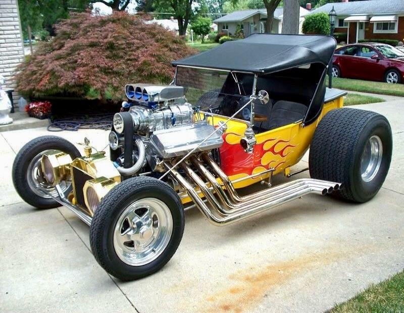A customized hot rod with a flame-painted body, exposed chrome engine with multiple exhaust headers, large rear tires, and a black soft top, parked in a suburban driveway.