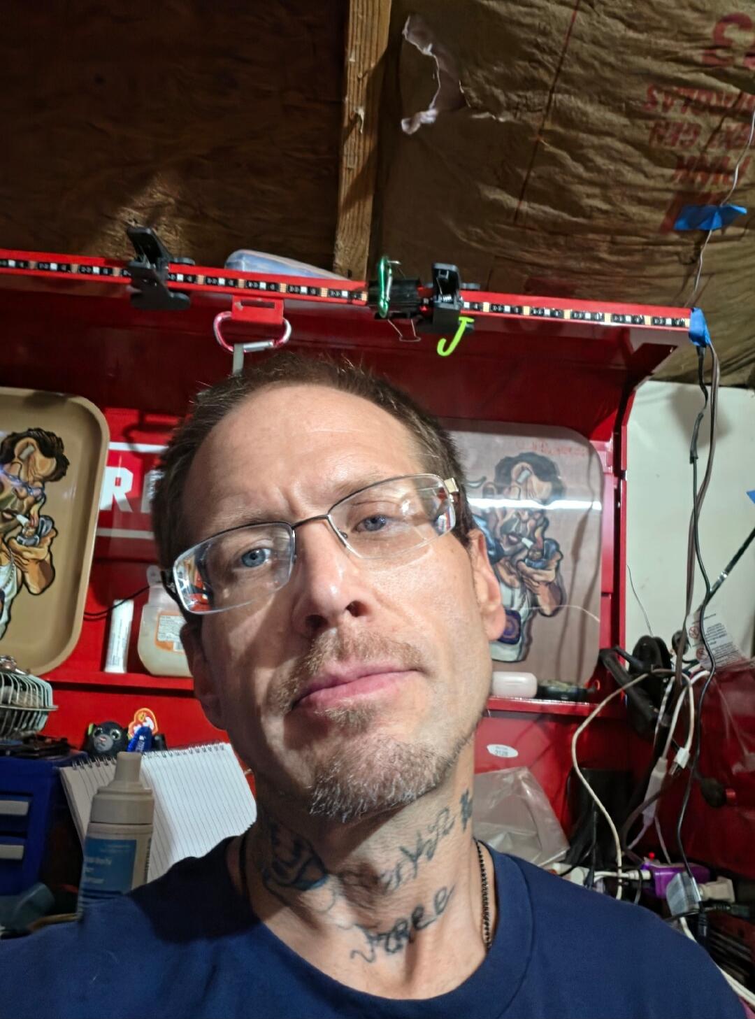 A man with glasses and neck tattoos takes a selfie in what appears to be a garage or workshop. In the background, there's a red toolbox with an 'R' visible, LED strip lights, and a tray with a cartoon drawing of a smoking character.
