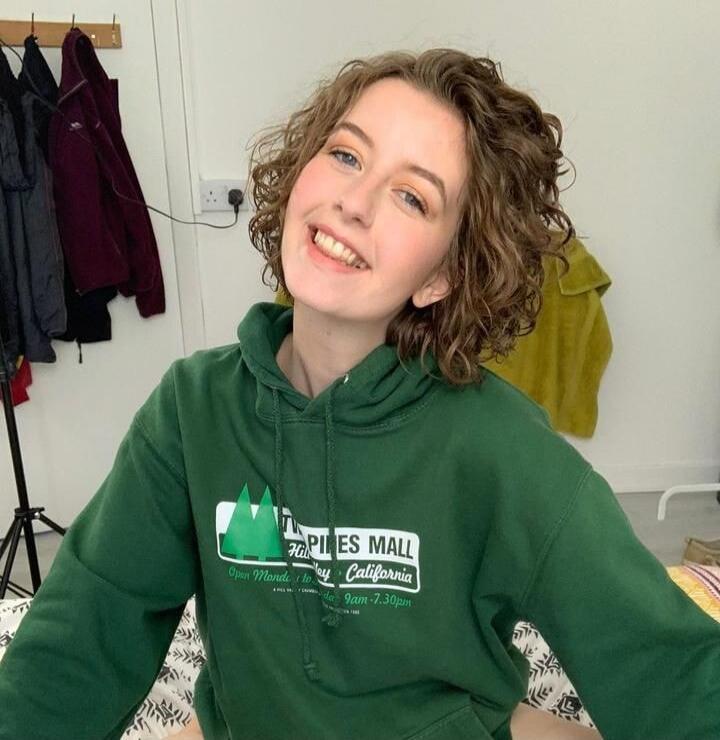 A young woman with curly hair is smiling brightly at the camera, wearing a green hoodie with 'TWO PINES MALL Hill Valley California Open Monday to Saturday 9am - 7.30pm' printed on it.
