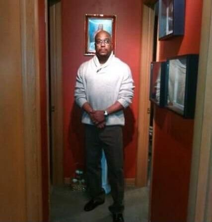 A man stands in a hallway, looking directly at the camera.