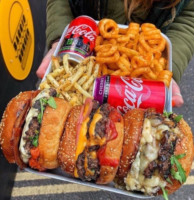 A tray filled with several gourmet burgers, two types of fries (curly and regular), and two cans of Coca-Cola (one classic, one Cherry Zero Sugar). Text on cans: 'Coca-Cola', 'CHERRY ZERO SUGAR', 'SUPER UPGRADE'. Text on sign: 'BABBIT BURGER'.