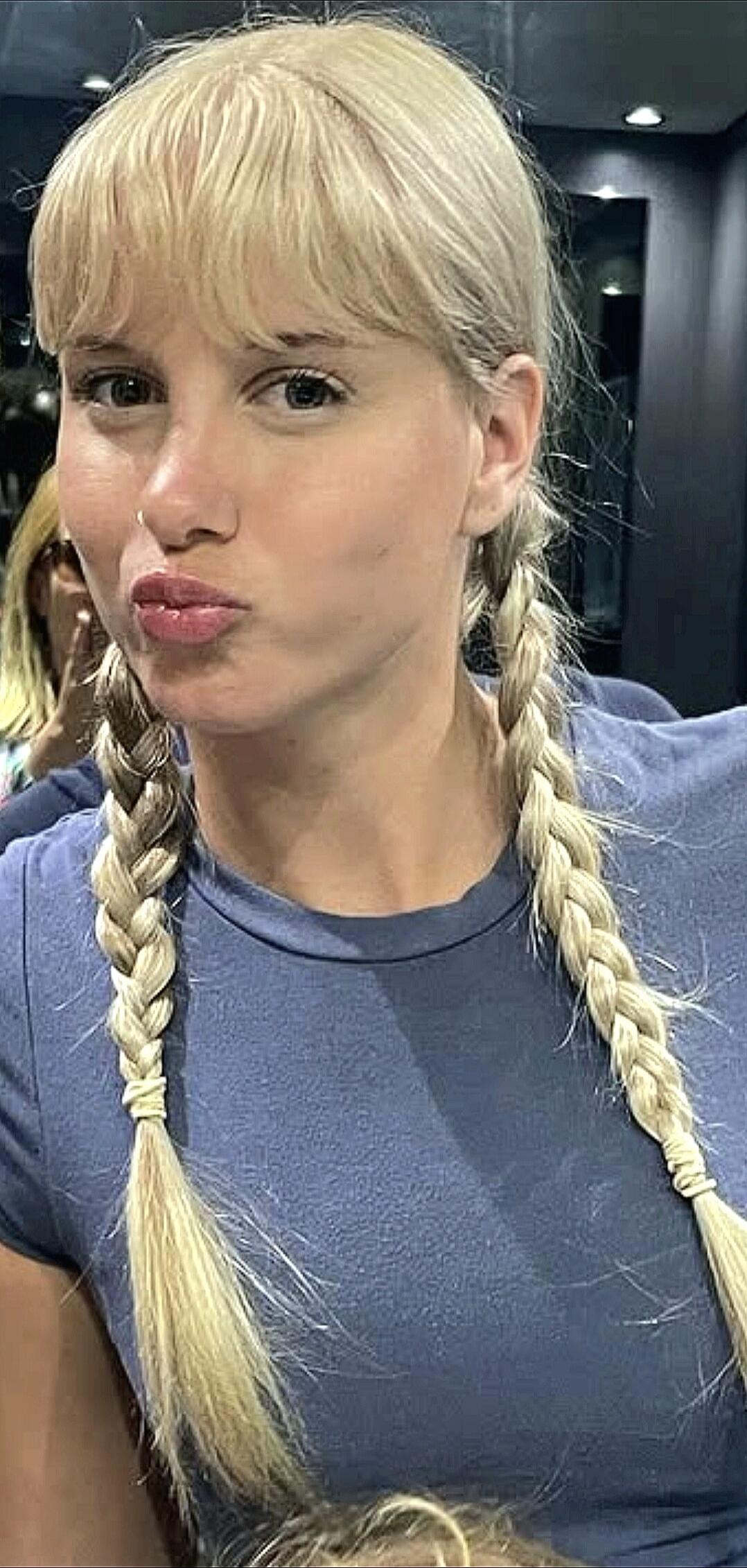 A blonde woman with pigtail braids and bangs, wearing a blue t-shirt, making a pouting or kissing face. She has a nose ring.