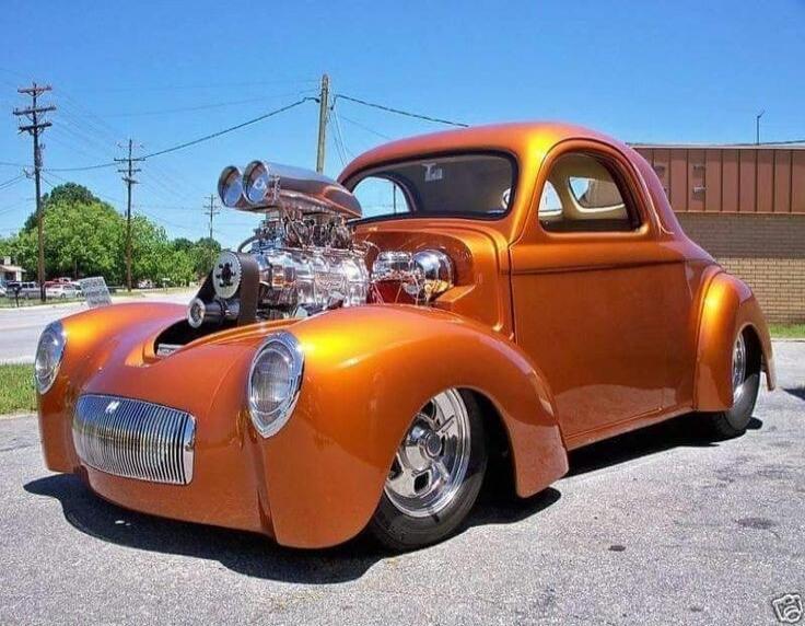 A custom orange hot rod car with a large exposed engine and supercharger.
