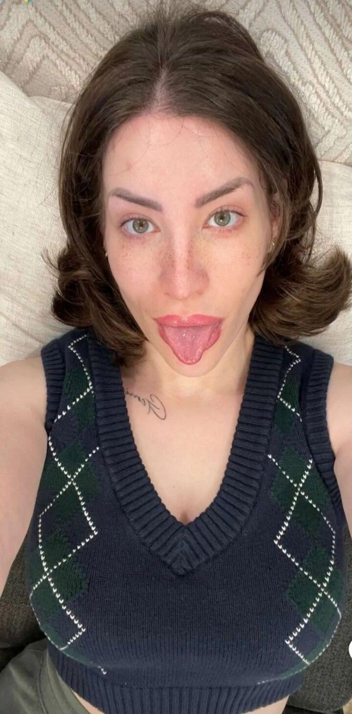 A woman with dark hair and freckles is looking at the camera and sticking her tongue out. She is wearing a dark blue argyle sweater vest.